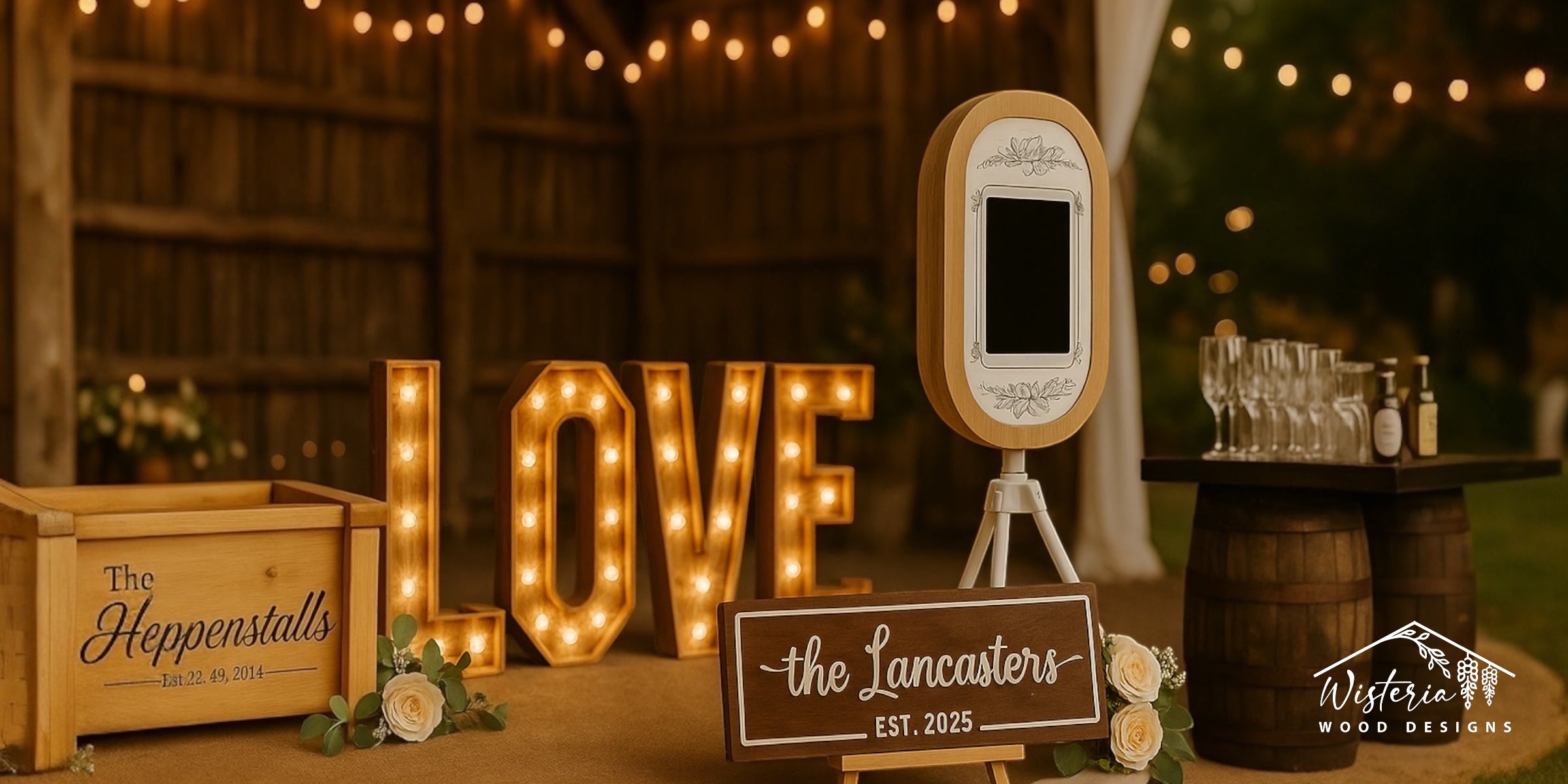 A full Wisteria Wood Designs wedding setup featuring a personalised keepsake box, illuminated LOVE letters, floral photo booth, drinks table, and a wooden name sign.
